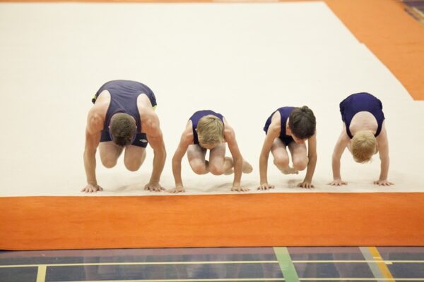 A coach and three boys in navy leotards crouched in their starting positions for a floor routine
