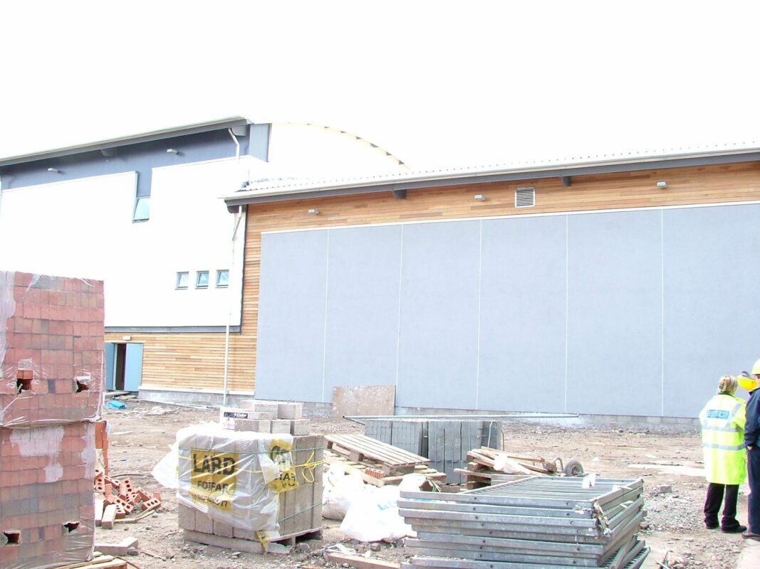 New building rising on a construction site, timber cladding and grey wall panels going up beside stacks of brick and timber.
