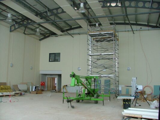 Inside the empty building shell, a tall scaffold tower reaches toward the curved steel roof structure.