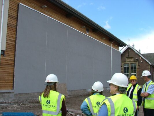 Visitors in Robertson Group hi-vis vests look up at the building's grey cladding, with the old school visible behind.