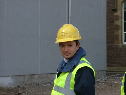 A visiting stakeholder in a yellow hard hat and hi-vis vest pauses for a photo on site.