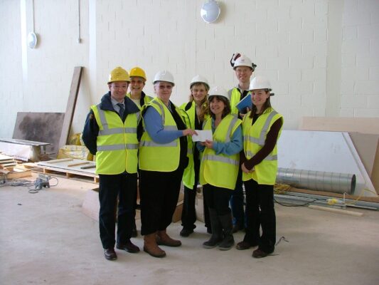 A group of seven people in hi-vis vests and hard hats smile as a funding cheque is handed over inside the new building.