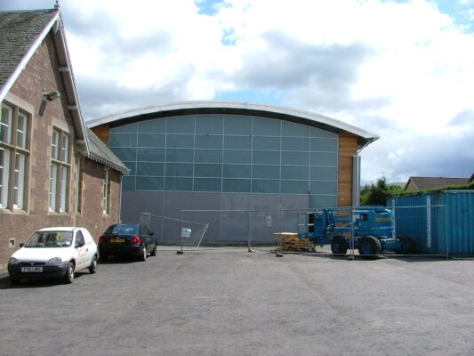 The finished building's curved roof and glass gable end, seen from the car park beside the old school.