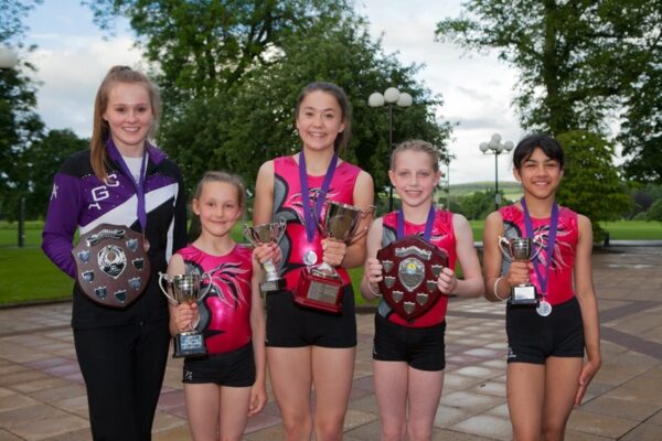 Five Auchterarder gymnasts in competition leotards holding trophies, shields and medals outdoors