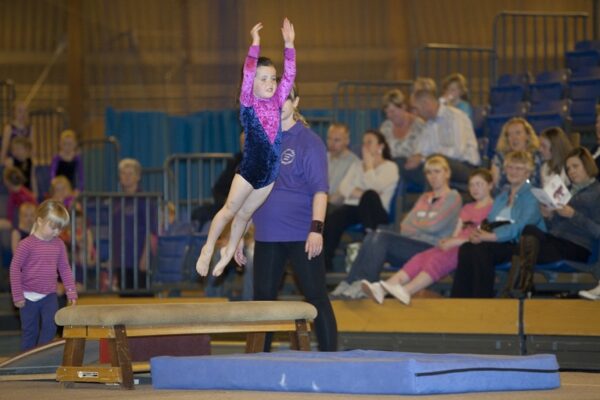 Young gymnast leaping from a padded training beam with a coach standing by at the 2012 club display