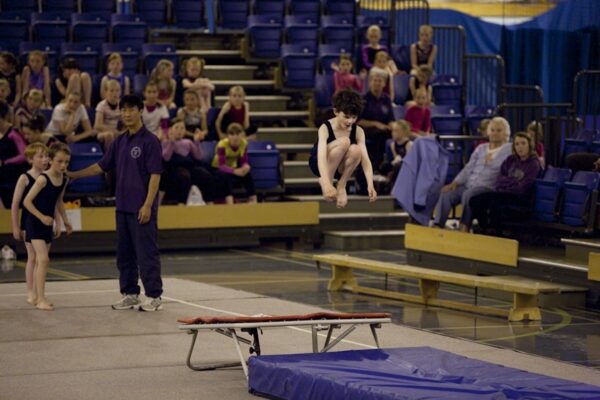 Boy performing a tucked jump from the trampette while a coach and waiting gymnasts look on at the 2012 club display