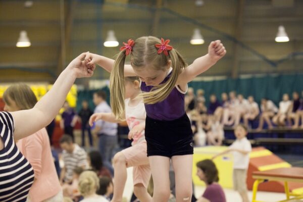 Toddler in a purple leotard jumping while holding an adult's hands at a pre-school gymnastics session