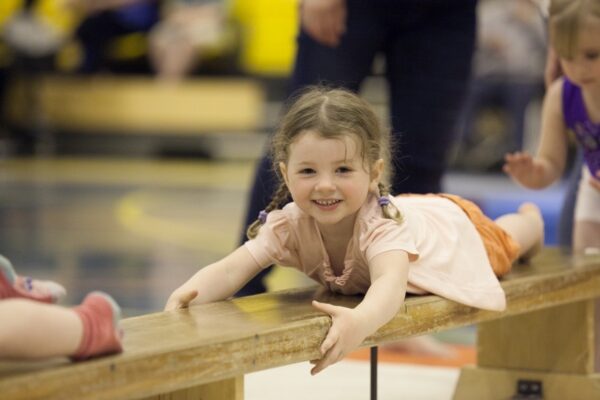 Smiling pre-school child lying across a low wooden bench during a toddler gymnastics session
