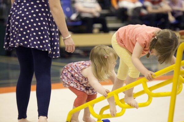 Two toddlers climbing a yellow frame with an adult supervising during a pre-school gymnastics session