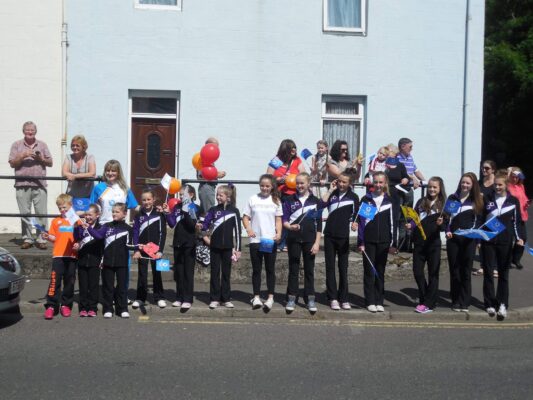 Auchterarder Gymnastics Club members in club tracksuits waving Scotland flags as they line the street for the Queen's Baton Relay